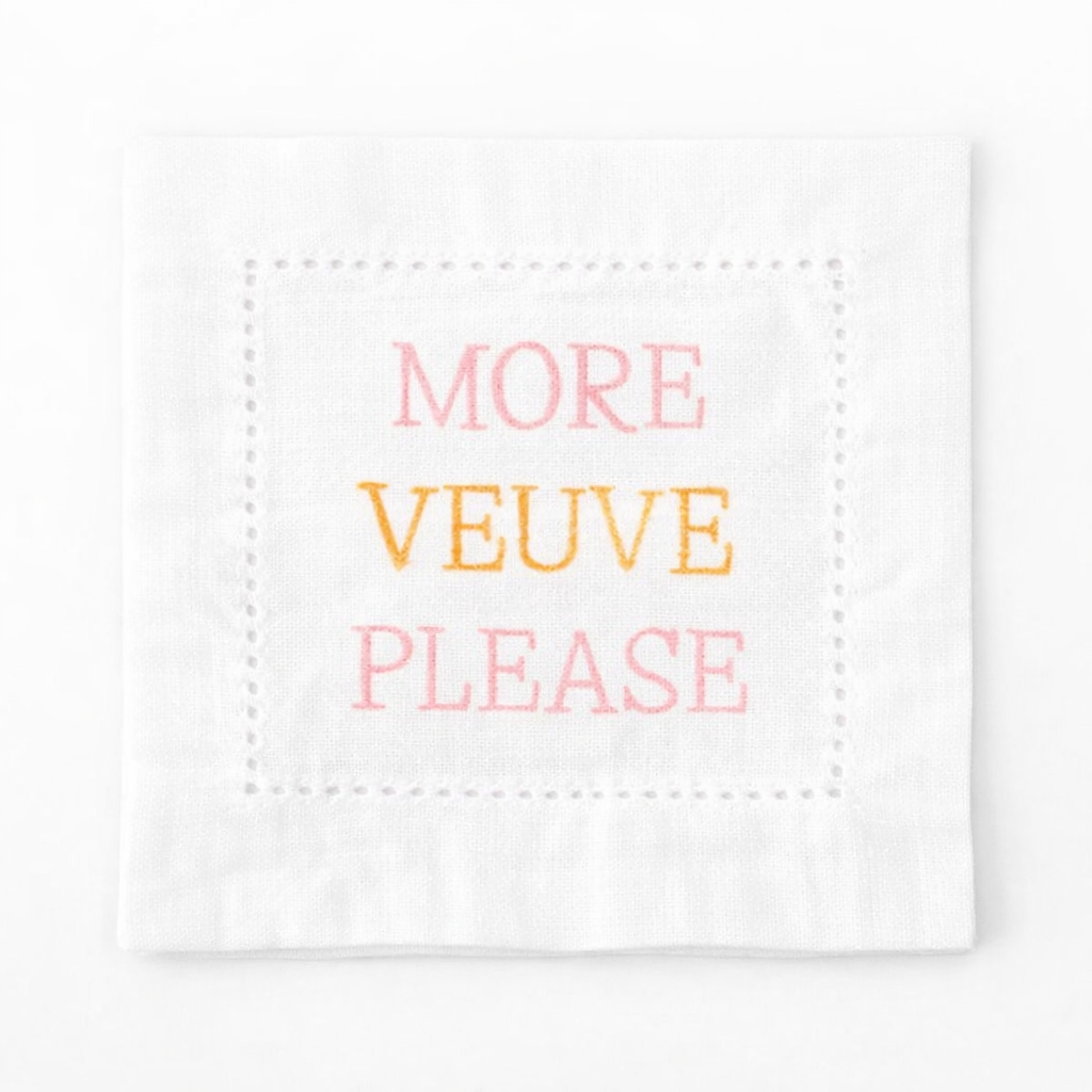 Embroidered white linen cocktail napkin featuring “More Veuve Please” phrase, styled with champagne glasses for bridal showers, girls’ brunches, and elegant entertaining.