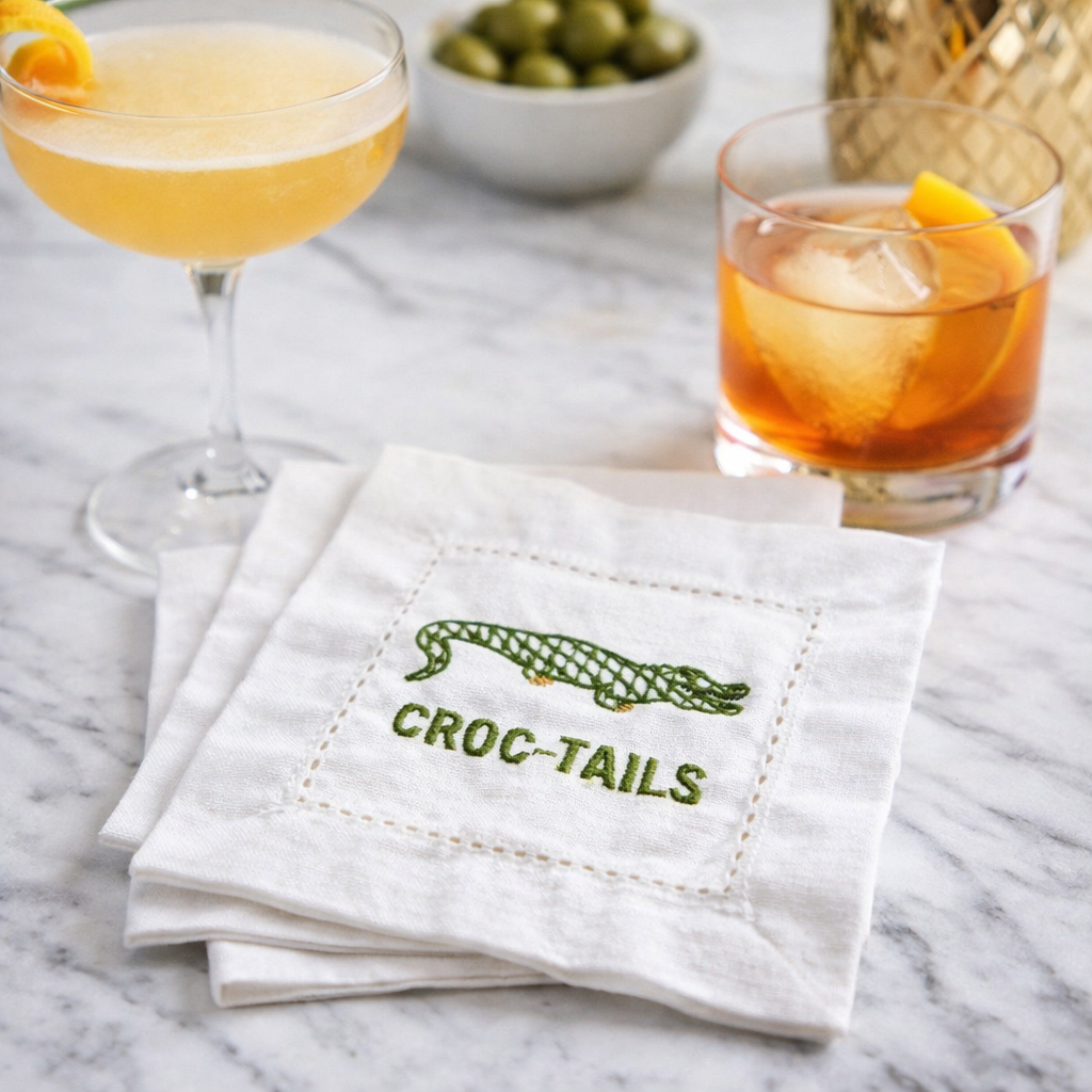 White embroidered cocktail napkin featuring a green alligator illustration with the text “Croc-Tails,” playful Southern cocktail party napkin with classic hemstitch border.