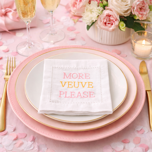 White embroidered cocktail napkin with “More Veuve Please” text in pink and gold thread, styled on a chic pink tablescape with champagne flutes, perfect for brunch, bridal showers, bachelorette parties, and entertaining.