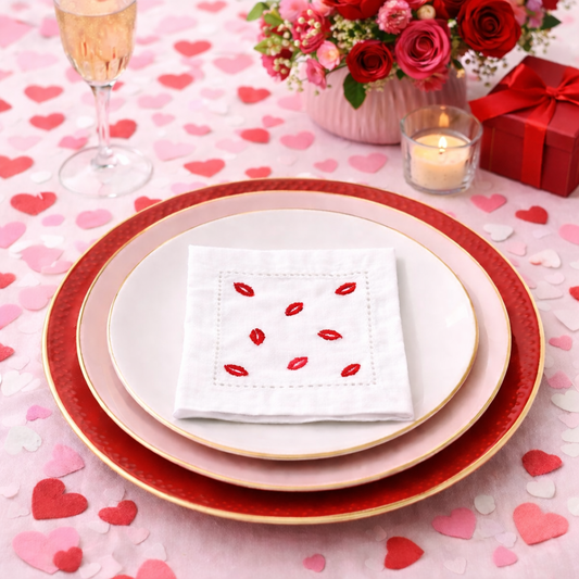 White embroidered cocktail napkin with red lip motif, styled on a romantic Valentine’s Day table setting with red and pink plates, perfect for entertaining, hostess gifts, and holiday décor.